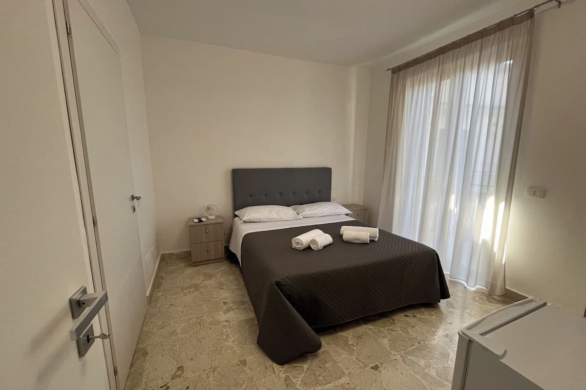 Ein Hotelzimmer mit Bett, Nachttisch, Lampe und einem Fenster mit Vorhängen.