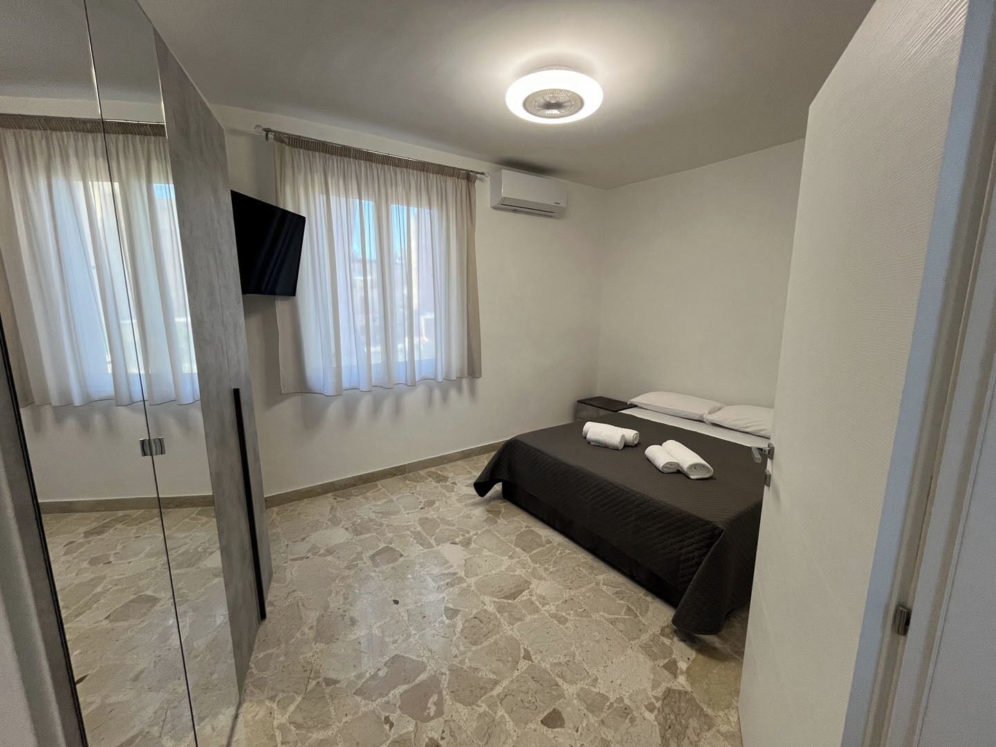 Un dormitorio con suelo de baldosas de piedra, una cama con ropa blanca, un televisor montado en la pared y una lámpara en el techo.