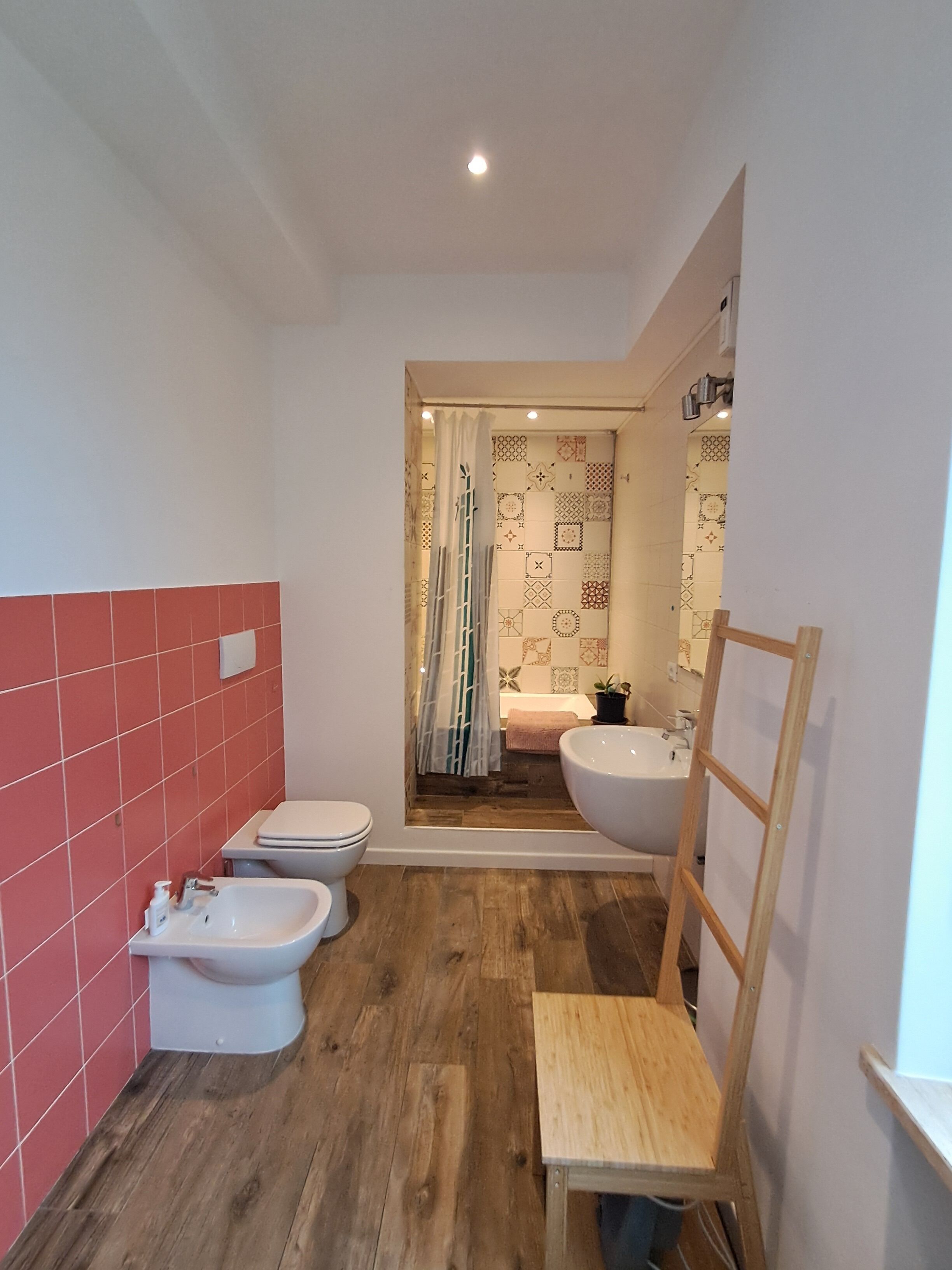 A bathroom with a wooden floor, a white sink, a bidet, and a toilet. There is a shower curtain with a geometric pattern.