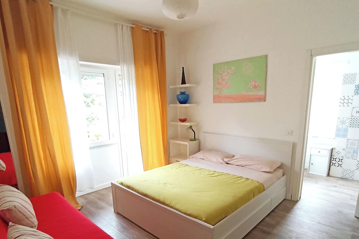 A bedroom with a bed, a red sofa, a window with curtains, a shelf with decorative items, and a painting on the wall.