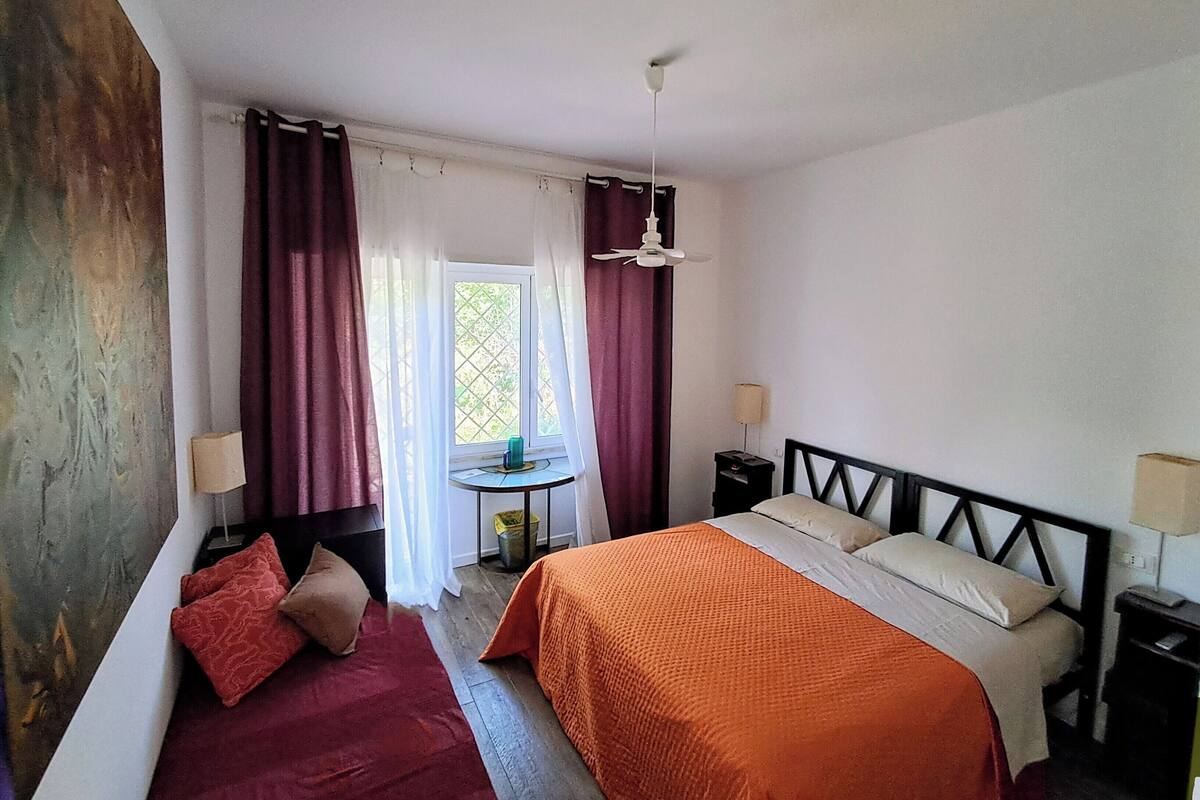 A bedroom with a bed, two bedside tables, a small table with a lamp, and a window with curtains.