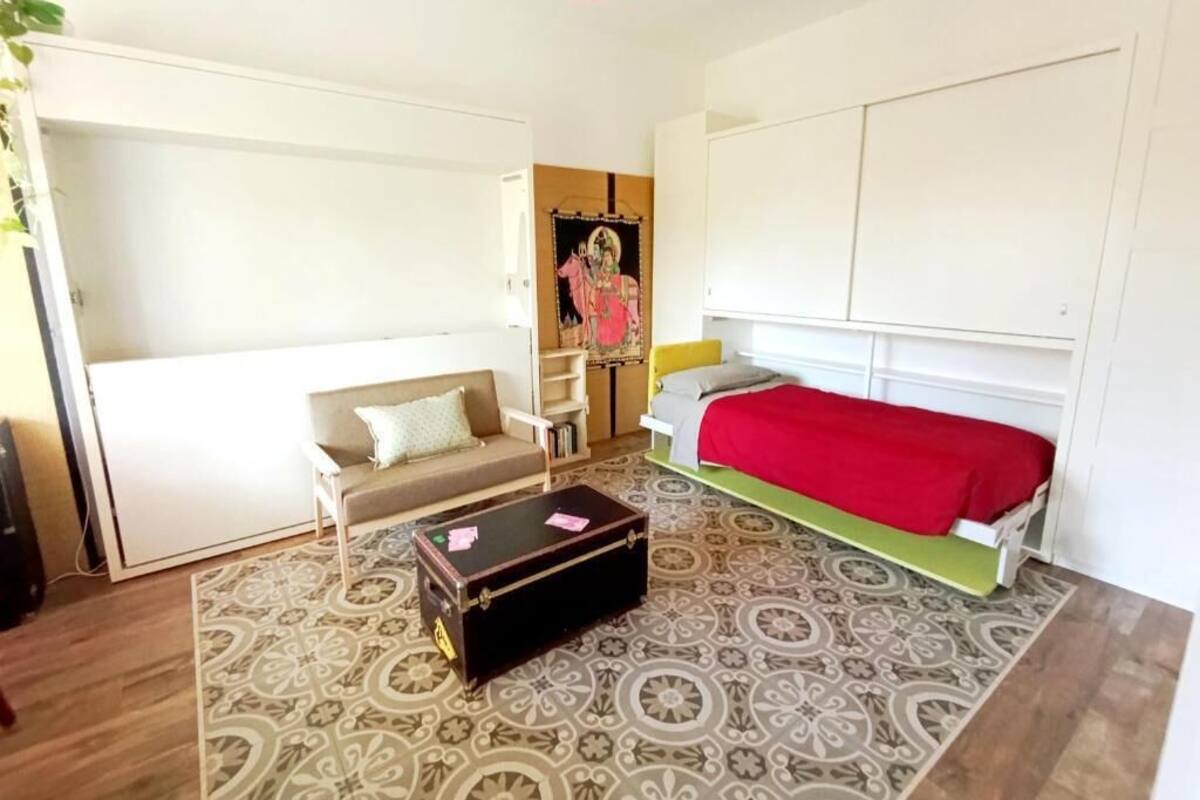 A room with a sofa, a bed with a red blanket, a patterned rug, and a wall with a framed picture.