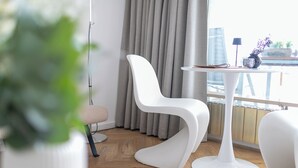 Une chambre moderne avec une chaise blanche, une petite table ronde et une vue sur le paysage urbain, visible à travers de grandes fenêtres.