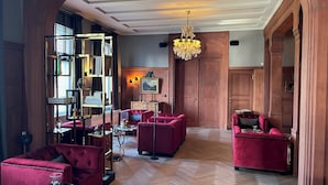 Een ruime woonkamer met een visgraat parketvloer, rode fluweelzachte banken, een houten boekenkast en een kroonluchter.