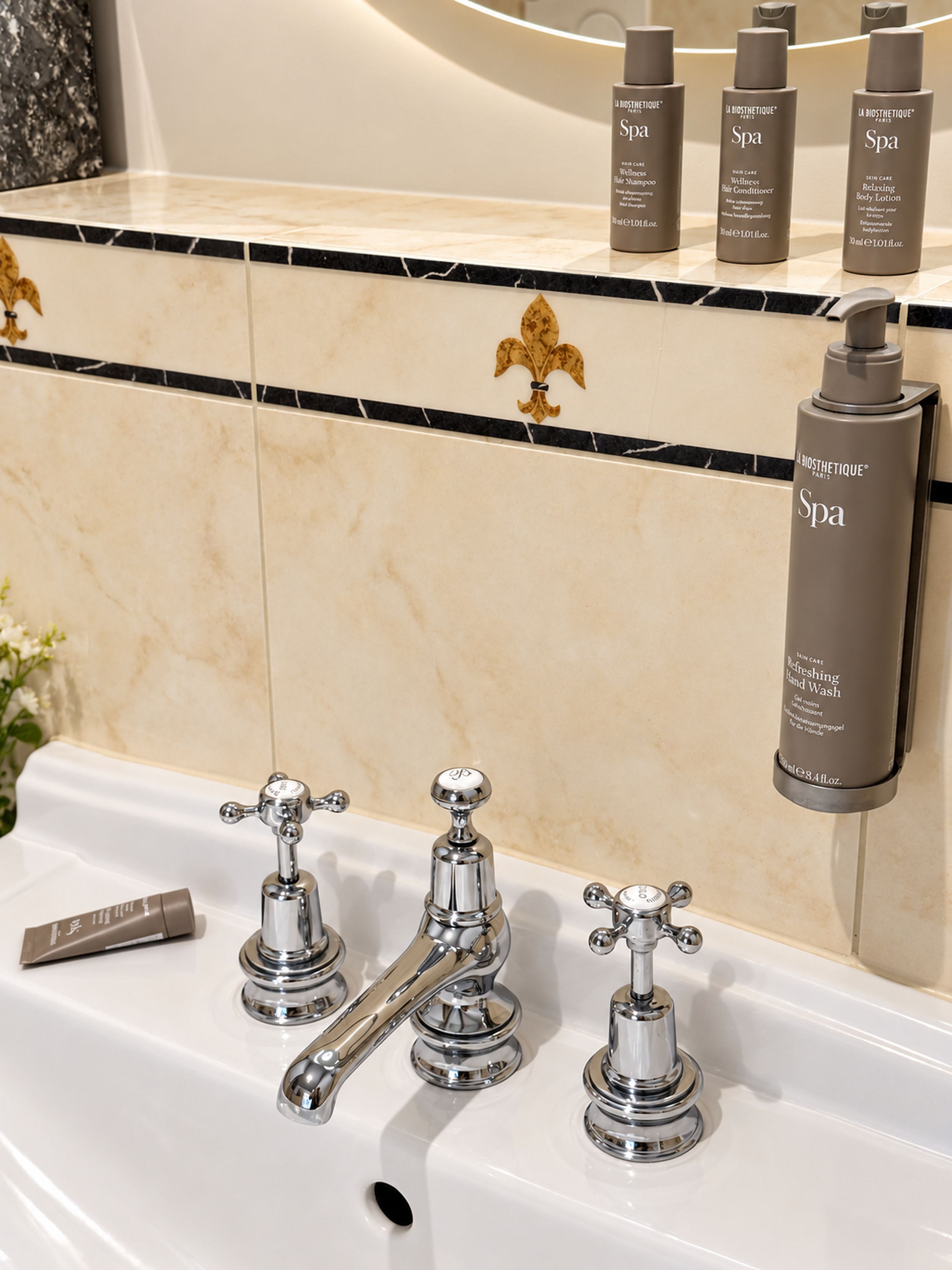 A bathroom with a marble countertop, a gold fleur-de-lis decoration, and three chrome faucets.