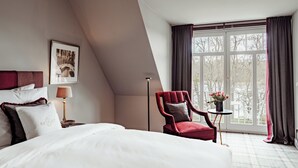 Hotelkamer met een groot bed, een rode fauteuil, een klein tafeltje met bloemen en een raam met gordijnen.