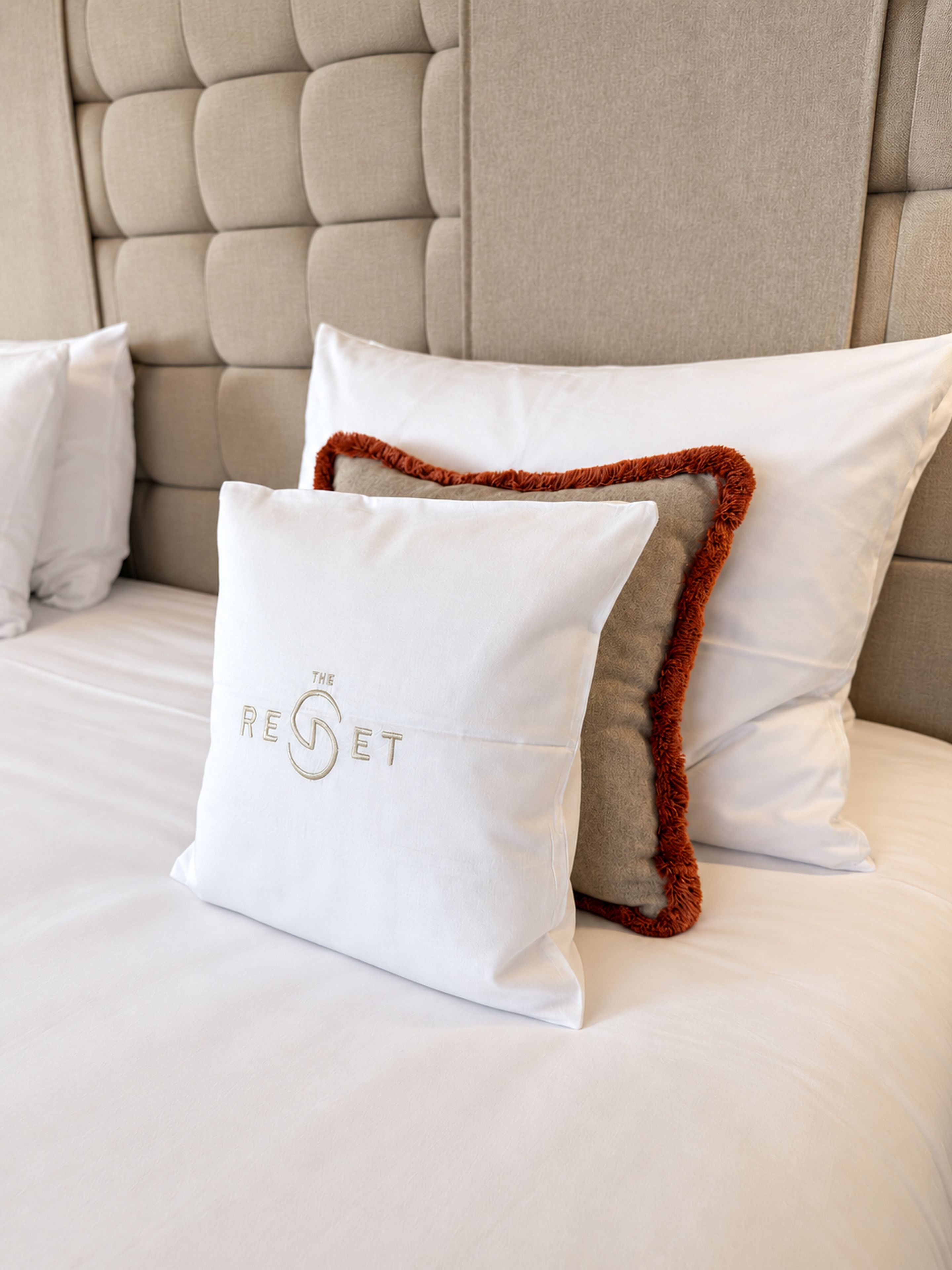 Una cama con cabecera capitonada, almohadas blancas y una almohada con texto bordado.