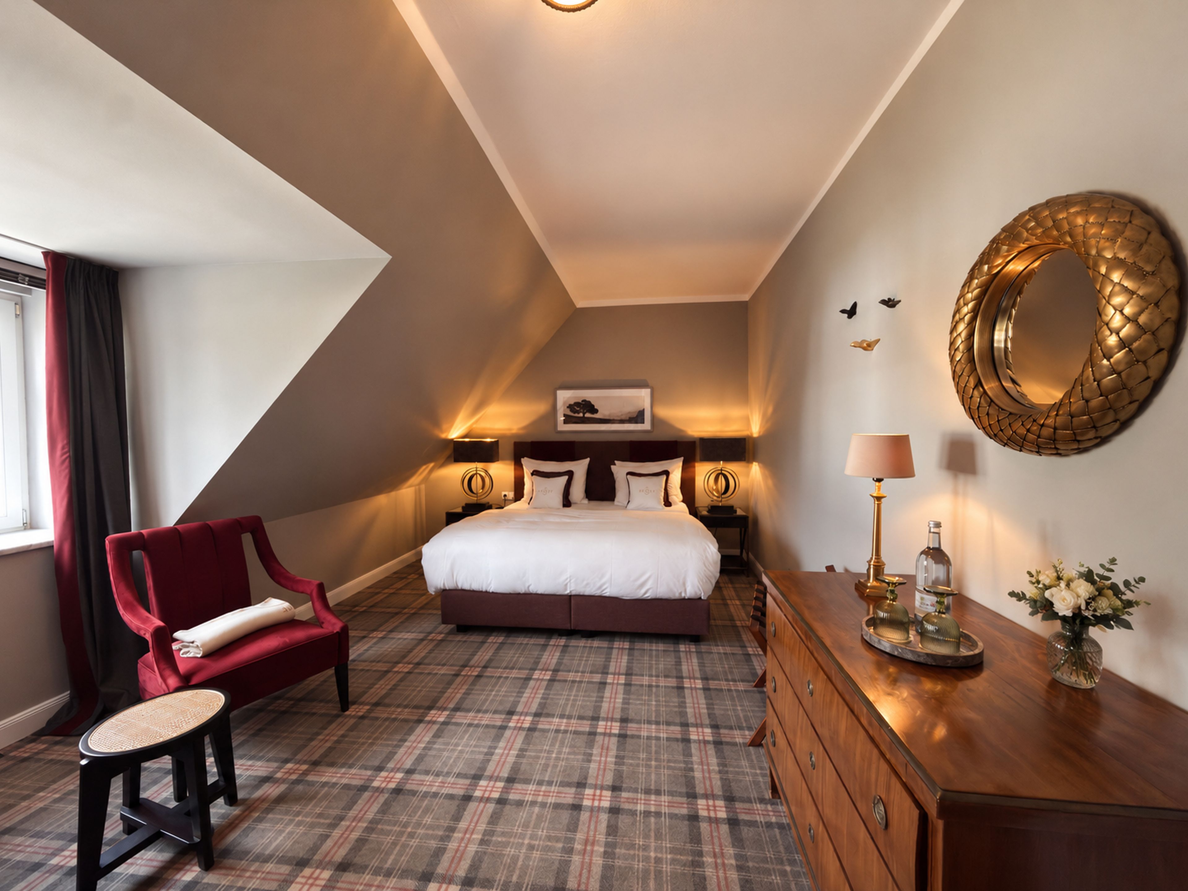 A hotel room with a bed, a red armchair, a small table, a dresser, and a mirror on the wall.