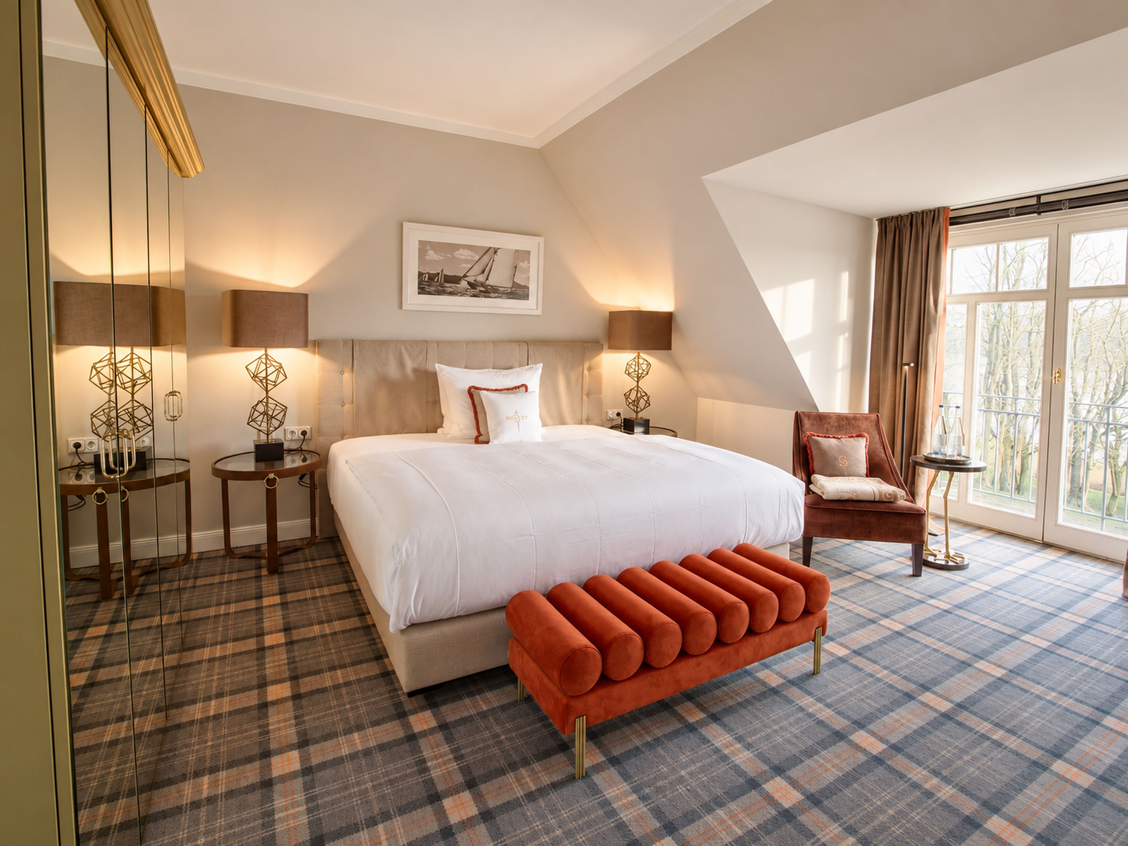 A hotel room with a large bed, a bench, a chair, and a large window with curtains.