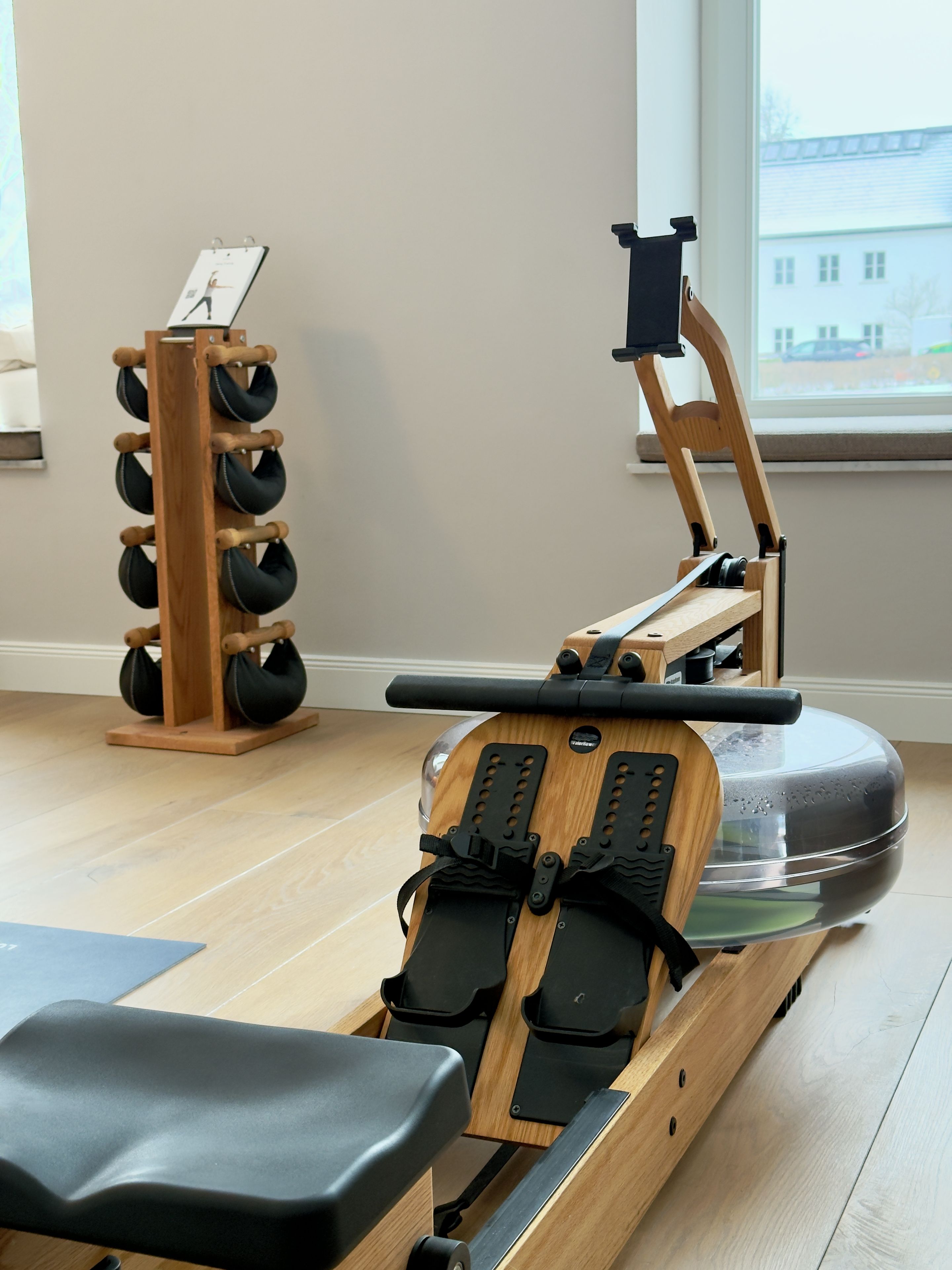 A rowing machine in a room with a wooden floor, a window with a view of buildings, and a wall-mounted rack with black bags.