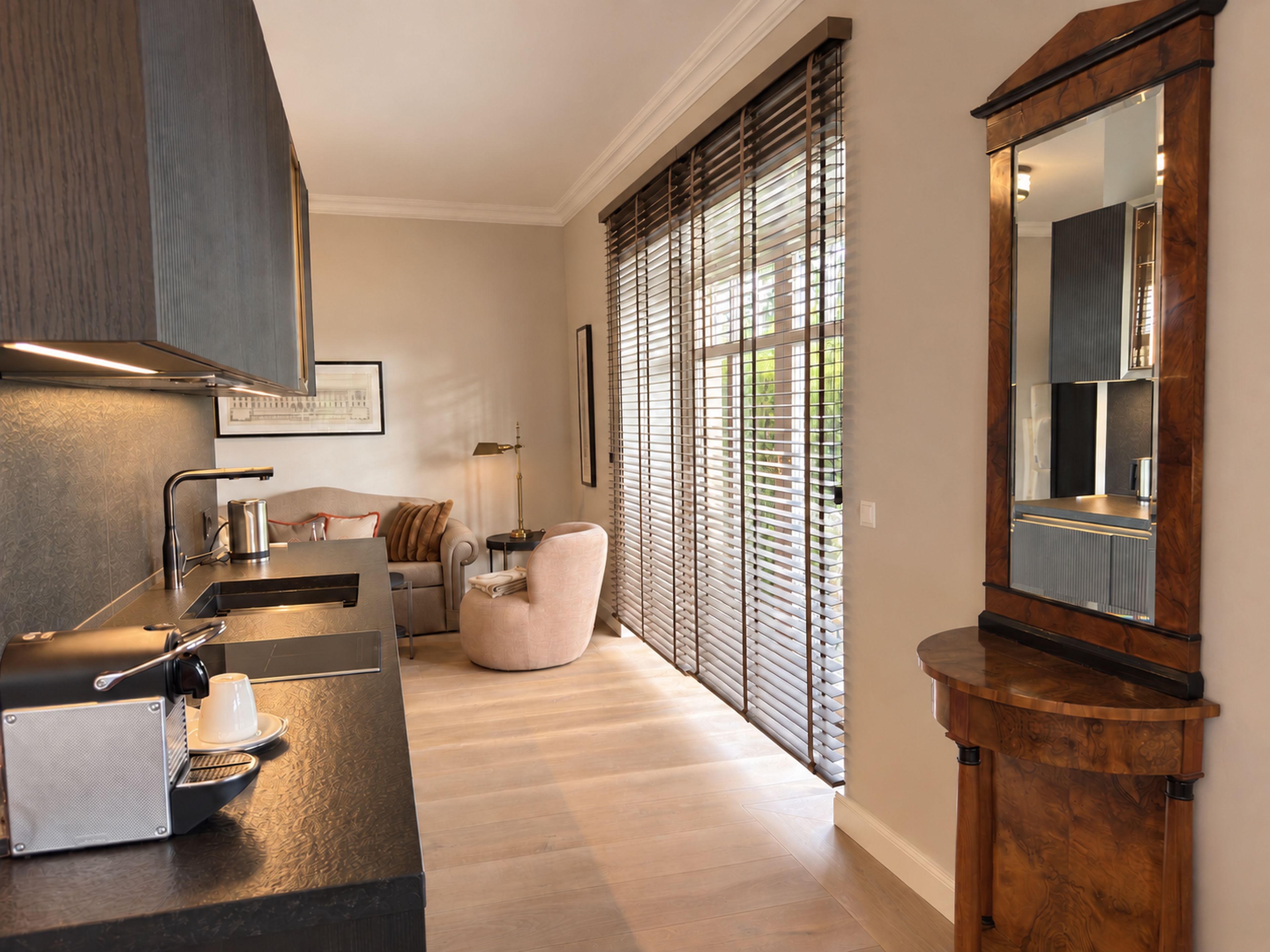 A modern kitchen with a built-in espresso machine, a seating area with a sofa and armchairs, and large windows with blinds.