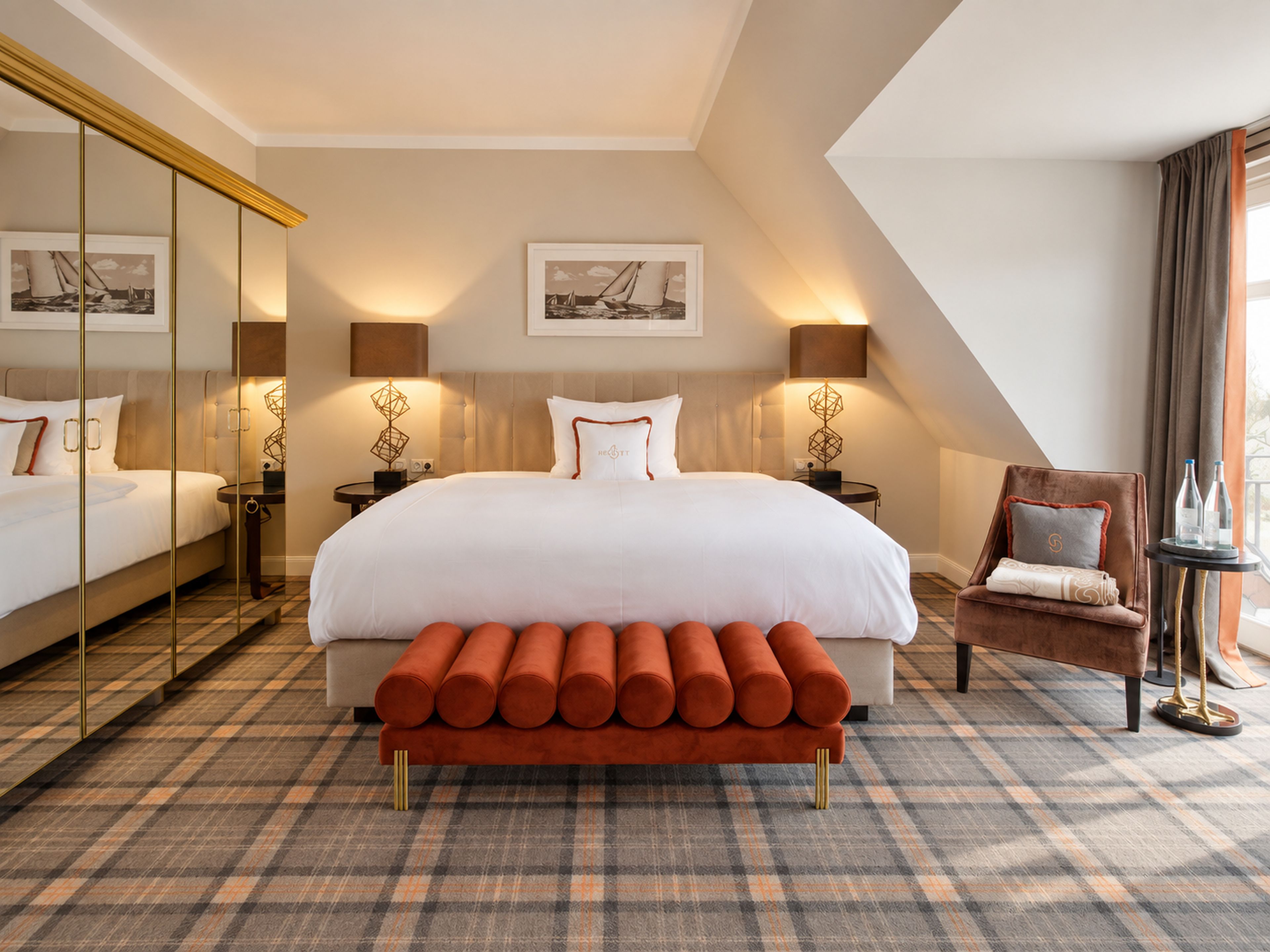 A hotel room with a large bed, a bench, a chair, and a small table with bottles.