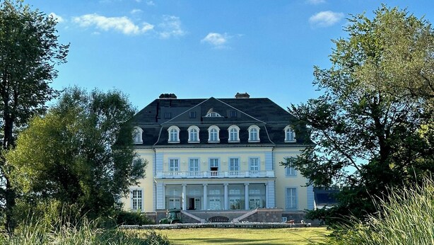 Een groot, historisch gebouw met een symmetrische gevel, een centrale ingang en een formele tuin op de voorgrond.