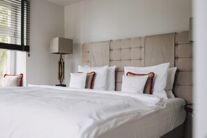 A neatly made bed with white linens and red-trimmed pillows, a beige upholstered headboard, and a bedside lamp. - THE RESET (Groß Plasten)