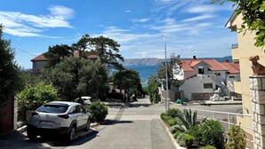 Une rue résidentielle avec une voiture garée, des maisons aux toits recouverts de tuiles rouges, et une vue sur la mer et les montagnes au loin.