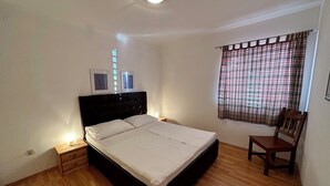 Une chambre à coucher avec un lit, un plancher en bois, une table de chevet, une chaise, une fenêtre avec un rideau à carreaux et un luminaire au plafond.