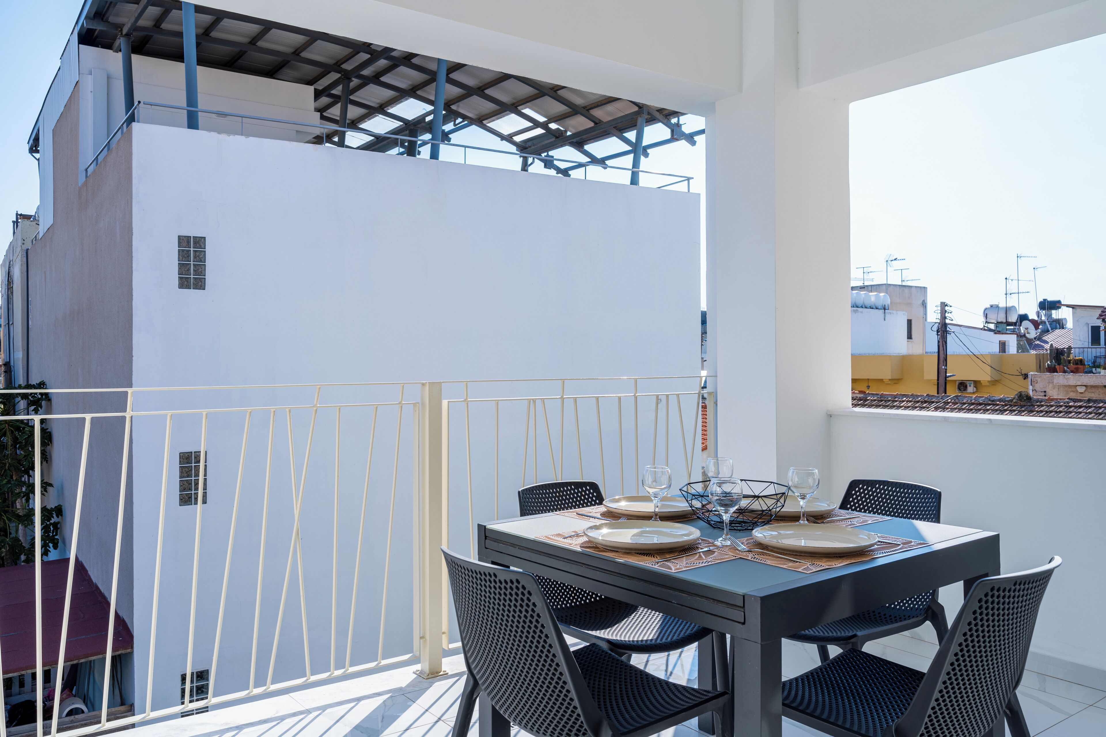 Un balcón con una mesa dispuesta para dos personas, con sillas de metal y superficie de vidrio.