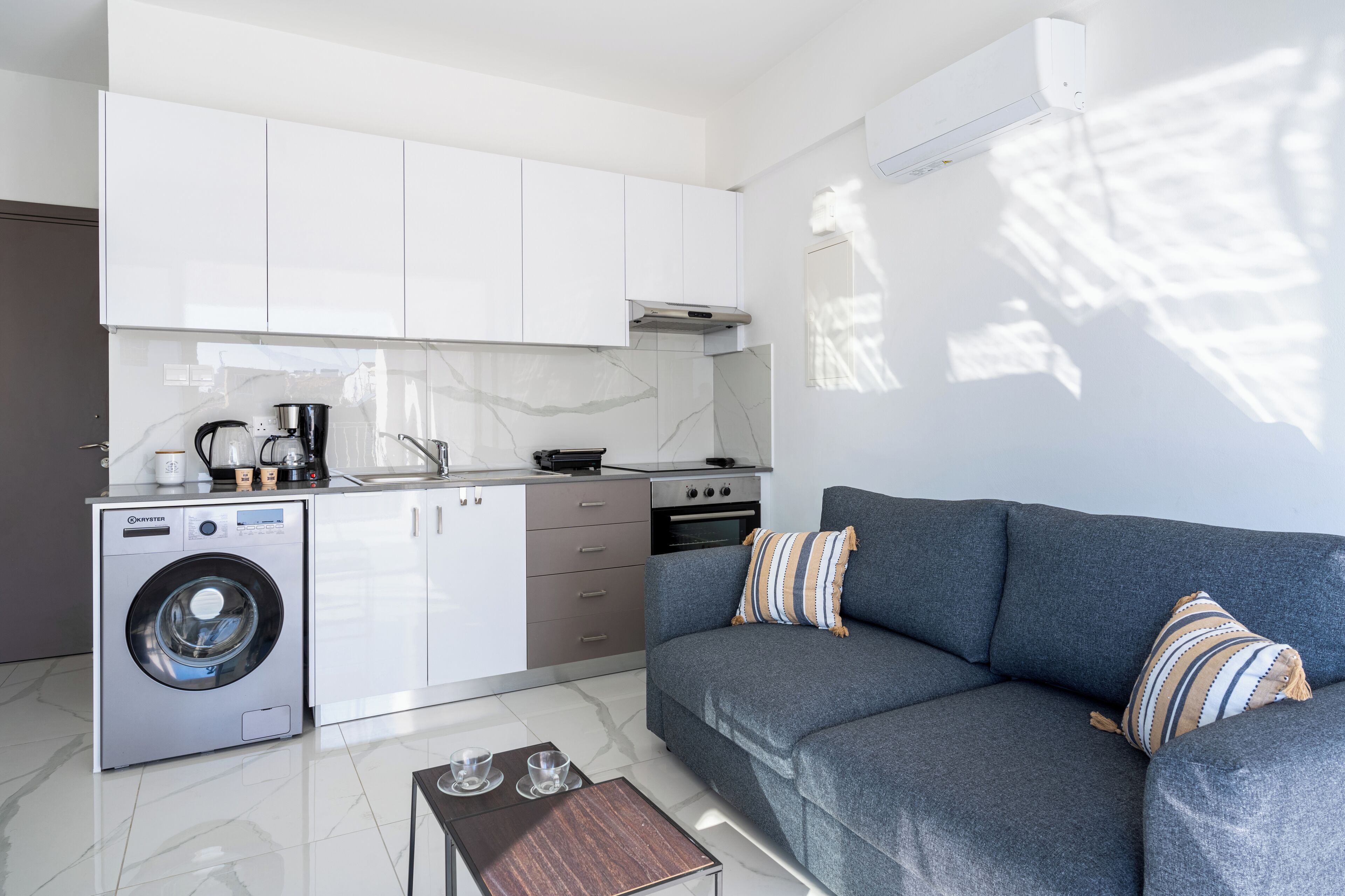 A modern kitchen with white cabinets, a built-in washing machine, and a grey sofa with striped pillows.
