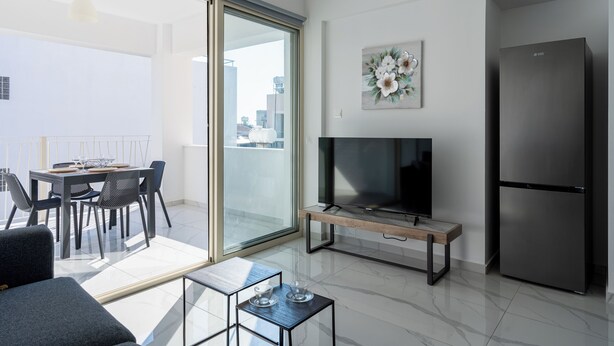 A modern living room with a flat-screen TV, a refrigerator, and a dining area with a table and chairs. - Rosemont Studio With Balcony in Larnaca (Larnaca)