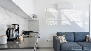 A modern kitchen with a coffee maker, kettle, and toaster. There is a grey sofa with striped pillows and a small table with a glass. - Rosemont Studio With Balcony in Larnaca (Larnaca)