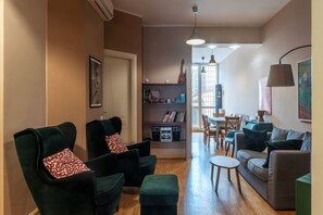 A living room with two green armchairs, a grey sofa, a small round table, a bookshelf with books and a guitar, and a dining area with chairs and a table.