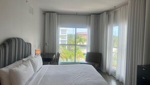 A hotel room with a large bed, a desk, and a chair. There is a window with curtains, and a view of palm trees and buildings outside.