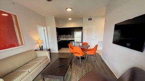 A modern living room with a sofa, coffee table, and orange chairs. There is a flat-screen TV mounted on the wall and a ceiling fan.