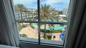 A hotel room with a view of a pool area, palm trees, and a building complex.