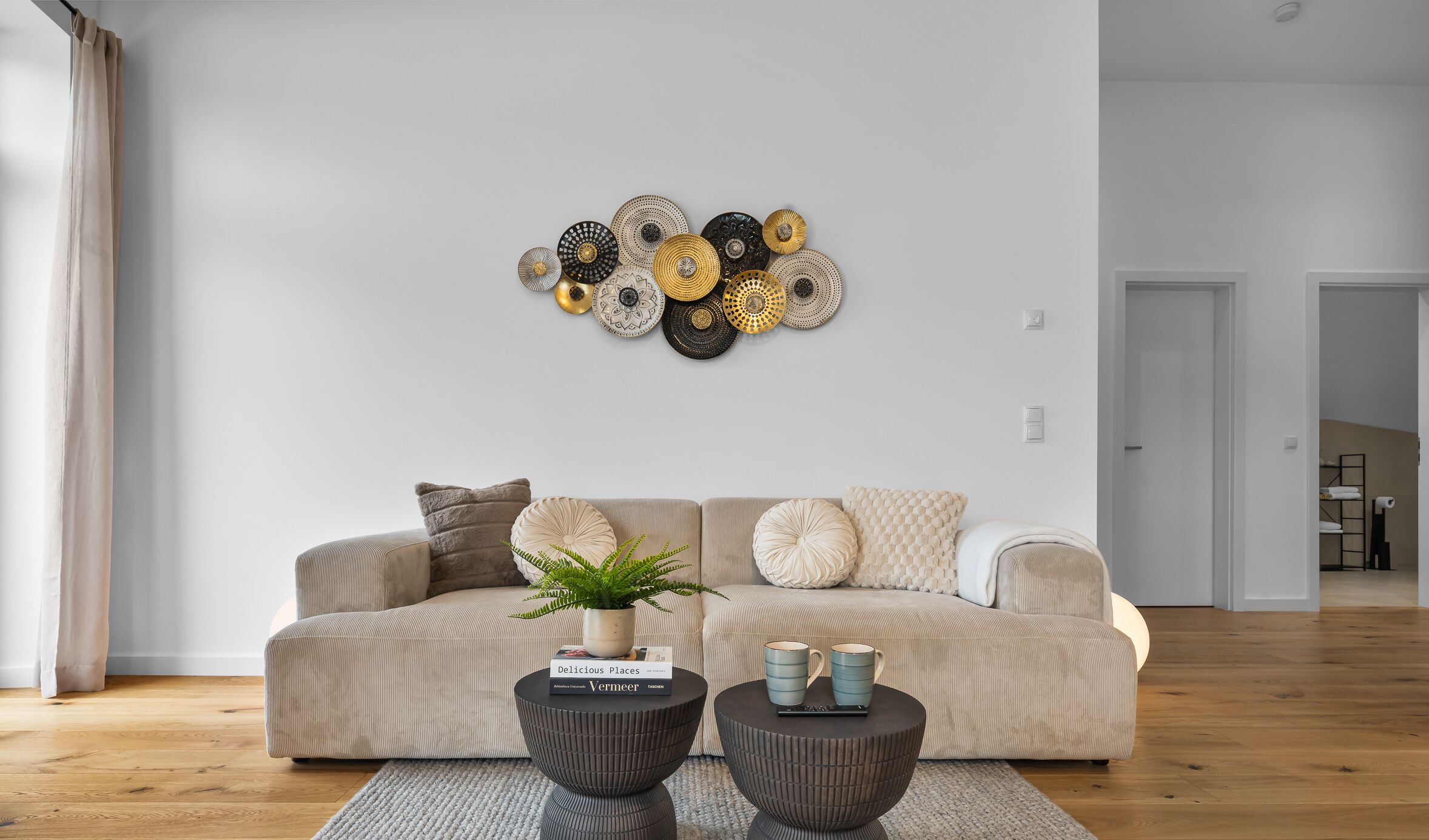 Ein modernes Wohnzimmer mit einem beigen Sofa, zwei runden Beistelltischen, einer Wanddekoration und einem Blumentopf.