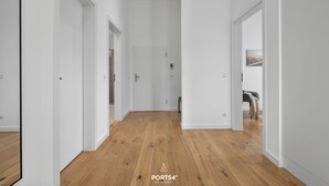 Un pasillo moderno y bien iluminado con suelo de madera y paredes blancas, que conduce a una habitación con una cama y un cuadro en la pared.