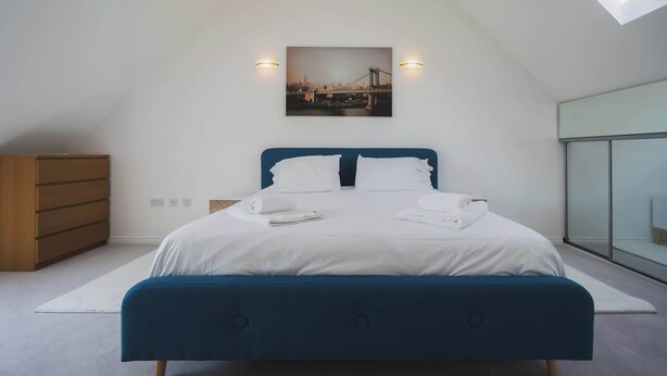 A modern bedroom with a large bed, a wooden dresser, and a cityscape painting on the wall. - Stylish 3-Bed Retreat with Garden and Parking (Woodford Halse)