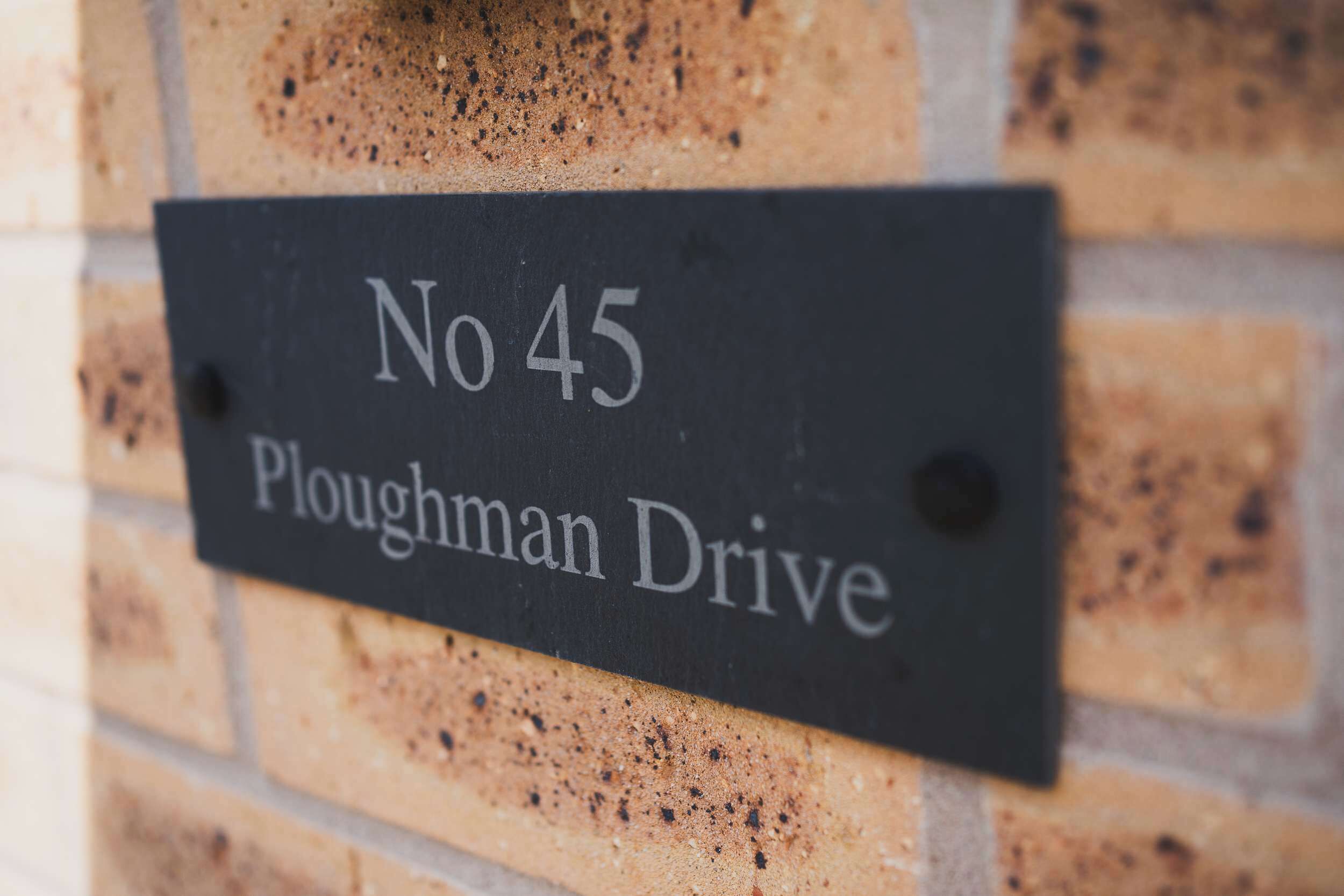 Ein schwarzes Schild mit weißer Schrift auf einer Ziegelwand, mit der Aufschrift 'No 45 Ploughman Drive'.