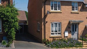 A two-story brick house with a sloped roof, a satellite dish, and a small garden with purple flowers. - Stylish 3-Bed Retreat with Garden and Parking (Woodford Halse)