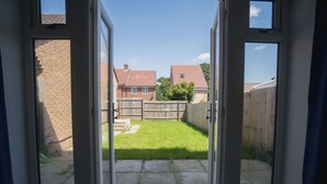 A view through a glass door into a backyard with a grassy lawn, a wooden fence, and a brick wall. - Stylish 3-Bed Retreat with Garden and Parking (Woodford Halse)