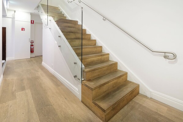 A staircase with wooden steps and a glass railing, leading to a room with a white wall and a fire extinguisher on the wall.