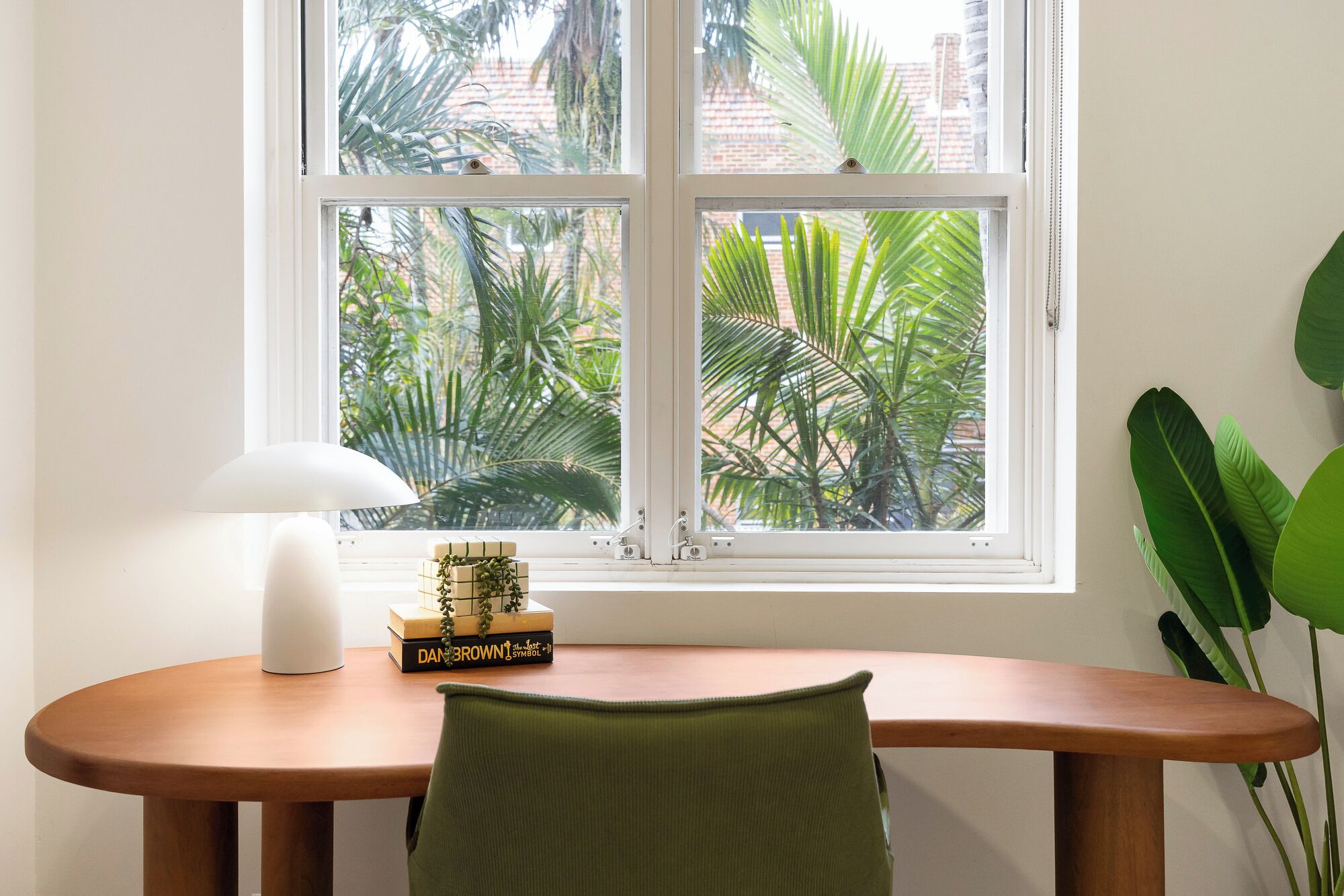 Una habitación con escritorio de madera, lámpara blanca y ventana con vista a palmeras.