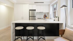 A modern kitchen with white cabinets, a black island with bar stools, and a window with blinds. - Urban Rest Double Bay Beach (Double Bay)