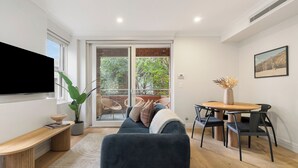 A modern living room with a flat-screen TV, a sofa, a dining table with chairs, a potted plant, and a view of the outdoors. - Urban Rest Double Bay Beach (Double Bay)
