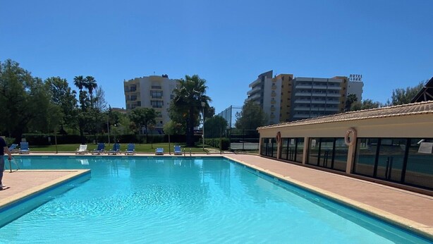 En swimmingpool med klart, blåt vand, omgivet af en flisbelagt terrasse og en bygning med teksten "HOTEL LURR" i baggrunden.