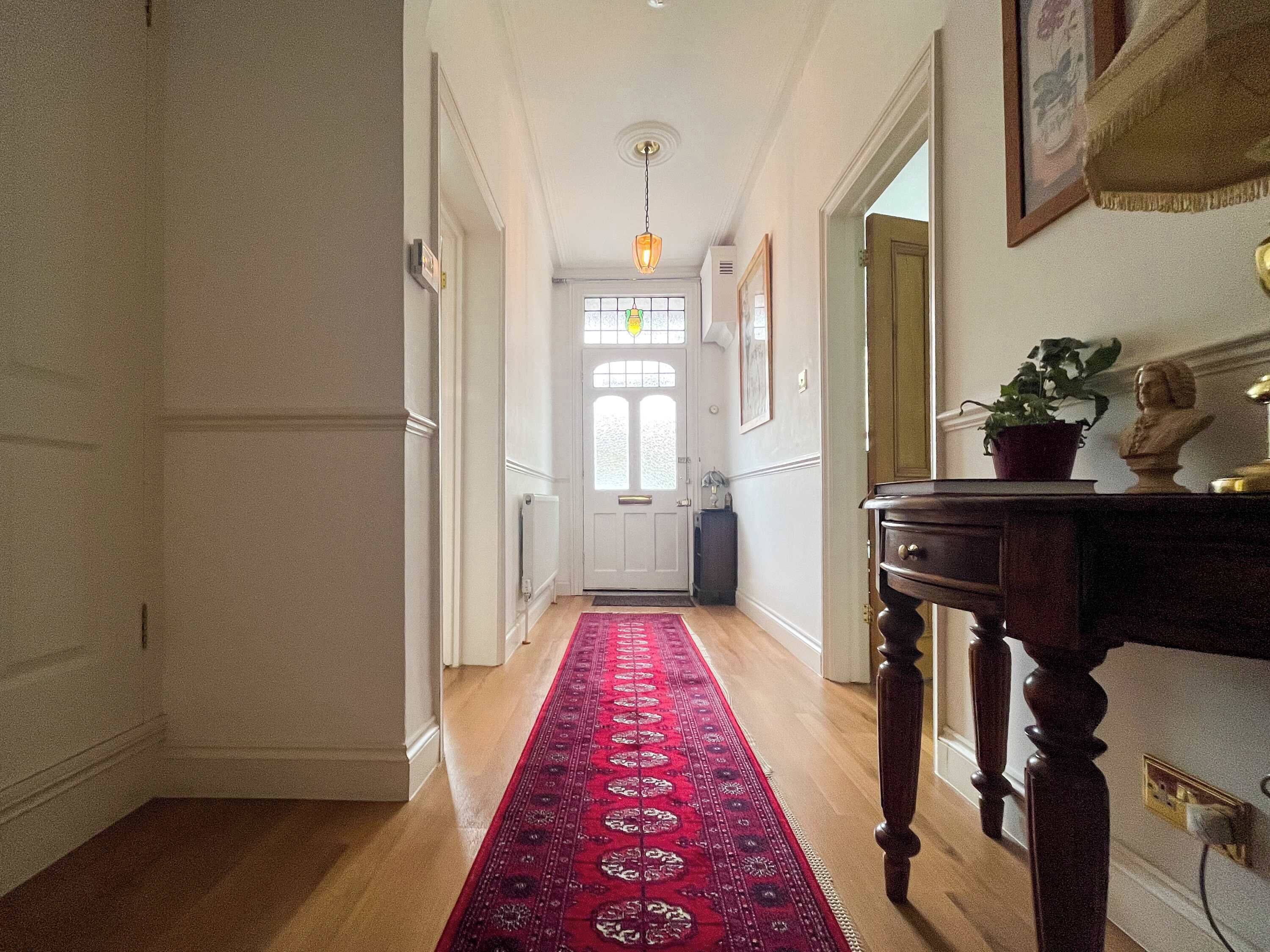 A hallway with a red carpet, wooden floor, a door with a glass panel, a wall-mounted lamp, and a bust on a table.