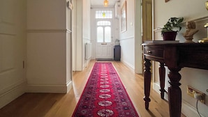 Un couloir avec un tapis rouge, un plancher en bois, une porte avec un panneau de verre, une lampe fixée au mur et un buste posé sur une table.