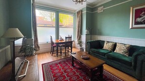 Un salon avec un canapé vert, une table basse en bois, un tapis à motifs rouges, une table à manger avec des chaises et une grande fenêtre avec des rideaux.