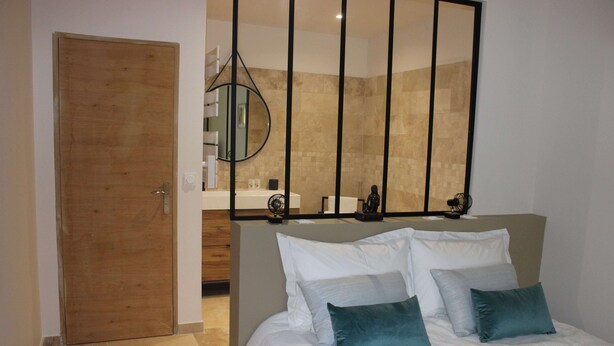 Une chambre avec une porte en bois, un lit avec une literie blanche et des coussins turquoise, un grand miroir, et une salle de bain avec une baignoire et un porte-serviettes.