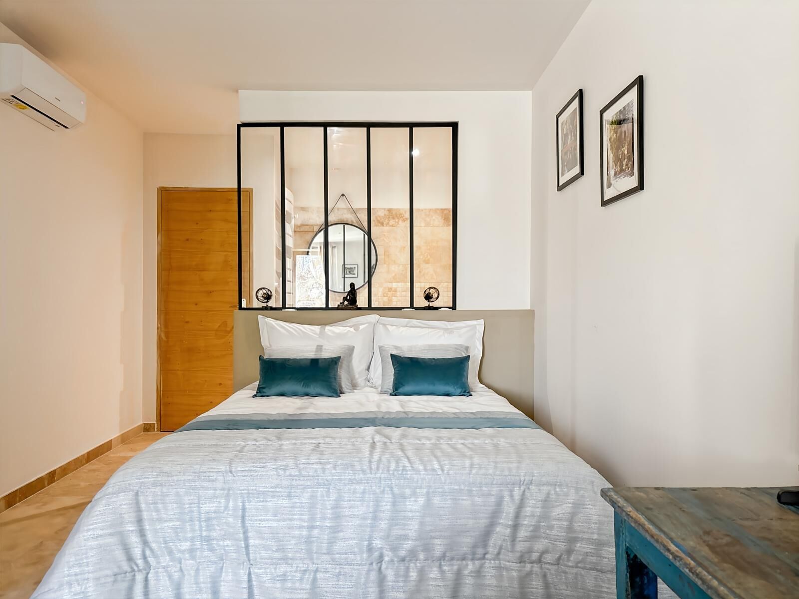 A bedroom with a bed, a wooden door, a wall-mounted mirror, and two framed pictures.