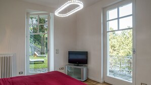 A bedroom with a bed, a television on a stand, and large windows with a view of greenery.