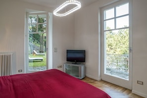 A bedroom with a bed, a television on a stand, and large windows with a view of greenery.
