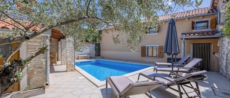 A swimming pool with lounge chairs, a stone wall, and a tiled patio.