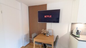 Een moderne kamer met een houten eettafel, twee grijze stoelen, een flatscreen televisie waarop 'NETFLIX' staat en een klein plankje met een apparaat en een ingekaderde welkomstkaart.