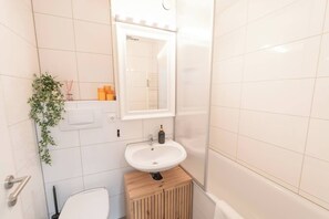 Ein kleines Badezimmer mit weißen Fliesen, einem Holzschrank, einem weißen Waschbecken, einem Spiegel, einer Toilette und einer Pflanze.