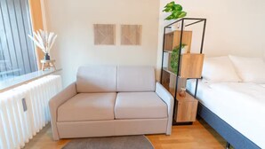 Een moderne woonkamer met een beige bank, een houten vloer, een witte radiator en een bed met witte lakens.